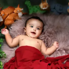 A happy baby lies on a soft fur rug, surrounded by greenery and woodland animal toys, smiling up at the camera.