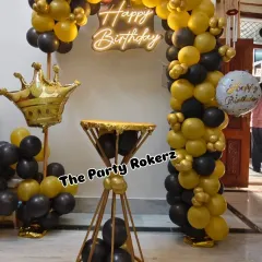 A stylish and modern birthday setup in black and gold. The decor includes a balloon arch, a neon sign, a diamond-shaped cake stand, and a crown foil balloon pillar.