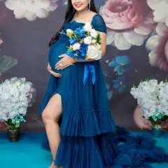 A vibrant and joyful solo portrait celebrating the final stages of pregnancy. The mom-to-be glows in a royal blue ruffled gown, complemented by a lush floral backdrop and a matching bouquet, creating a rich, colorful scene.