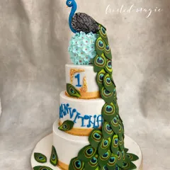 A majestic peacock-themed cake for a first birthday celebration. The peacock's body is sculpted and its cascading tail is made of dozens of individually crafted and painted feathers.