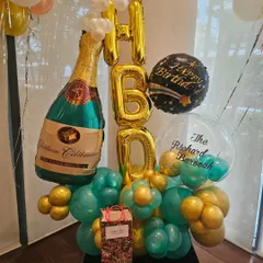 A celebratory balloon bouquet perfect for an adult's birthday. This arrangement features a large champagne bottle foil balloon, 'HBD' letters, and a mix of teal and gold balloons.