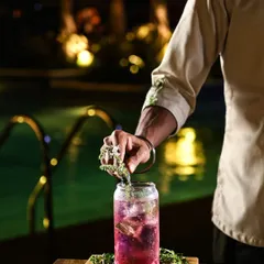 As night falls, our poolside bar Asri comes alive. A bartender carefully garnishes a vibrant, purple-hued cocktail, showcasing the artistry and attention to detail that goes into every drink.