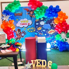 A powerful superhero-themed party for Ved's 6th birthday. This setup includes a comic-book style backdrop with Superman and Iron Man, themed plinths, and marquee letters.