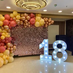 Celebrate turning 18 in style with this vibrant setup. A rose gold sequin wall provides the sparkle, framed by a warm-toned balloon garland and lit by large '18' marquee numbers, setting the stage for an unforgettable night.