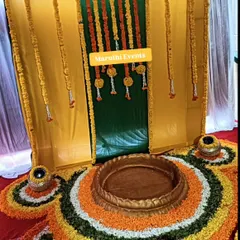 A traditional setup for a havan or puja, featuring a backdrop of yellow and green drapes with marigold garlands. The floor is covered with a floral rangoli, creating a sacred space for rituals.