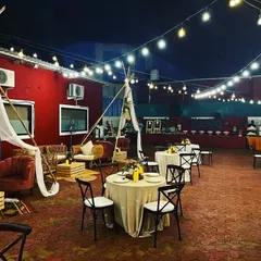 A rustic and inviting outdoor event setup. The space features bohemian-style lounge seating with teepees, round dining tables, and a buffet station in the background, all under a canopy of warm string lights.
