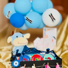 Welcoming a baby boy with this essentials hamper. It includes a soft cow plush toy, colorful rattles, diapers, and adorable printed bibs, all arranged in a handsome black crate decorated with a 'Just Arrived' balloon bouquet.
