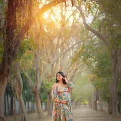 A stunning wide shot in a tree-lined path. The mother-to-be in a floral gown is perfectly framed by nature, with the golden sun creating a heavenly glow.
