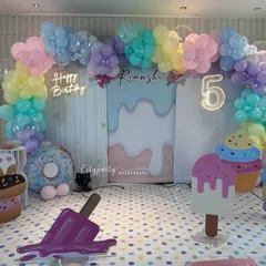 A sweet and colorful candy theme for a 5th birthday party. The setup includes a pastel rainbow balloon arch, a 'Happy Birthday' neon sign, and large props shaped like ice cream, donuts, and marshmallows.