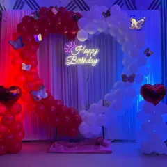 A romantic heart-shaped balloon arch for a birthday celebration. This design uses red and white balloons, butterfly accents, and a 'Happy Birthday' neon sign.