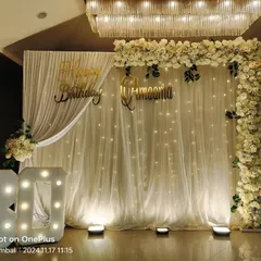 An exquisite all-white floral decoration for an 80th birthday celebration. The setup includes a white curtain backdrop with fairy lights, an L-shaped floral frame, and large '80' marquee numbers.