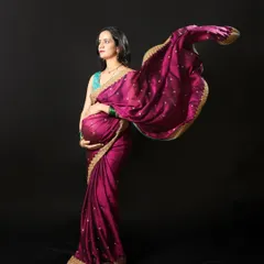 A dramatic and elegant maternity photo featuring a flowing magenta saree against a dark backdrop. This style of shot emphasizes the beautiful silhouette and the rich colors of the traditional fabric.