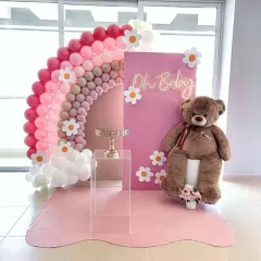 A fun and sweet baby shower backdrop featuring a rainbow balloon arch made of pink and neutral tones. The design is accented with cute daisy cutouts, an 'Oh Baby' neon sign, and a friendly teddy bear.