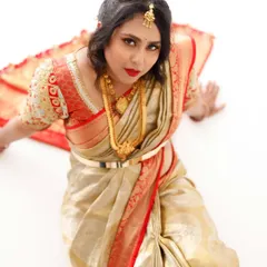 A high-angle shot from a bridal photoshoot. The client is looking up, showcasing the dramatic eye makeup and bold red lip that contrasts beautifully with her gold and red saree.