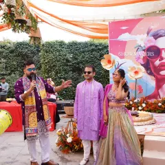 The happy couple on stage with the emcee, enjoying the festivities of their vibrant orange-themed Haldi function.