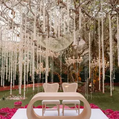 A modern and romantic mandap setup under a canopy of hanging floral strings and chandeliers. The unique, curved wooden table and minimalist chairs create a chic and intimate space for the wedding ceremony.