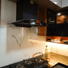 A close-up of a modern kitchen featuring a sleek black chimney against a white marble-veined backsplash. The under-cabinet lighting illuminates the granite countertop, highlighting the clean lines of the gas hob and the overall minimalist design.