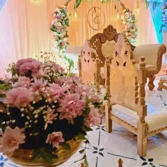 A detailed shot of the seating area for an engagement ceremony. The setup includes traditional wooden chairs for the rituals, placed in front of the main stage, with brass flower vases and lanterns adding to the decor.