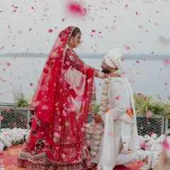 A romantic varmala moment with a shower of pink rose petals. The bride is in a stunning red lehenga, and the decor is set against a serene water body, making the entire scene feel like a beautiful painting.