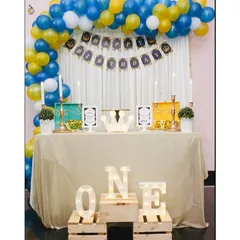 A royal prince theme for a first birthday party. The decor includes a blue, yellow, and white balloon arch, a personalized birthday banner, and light up 'ONE' letters on rustic wooden crates.