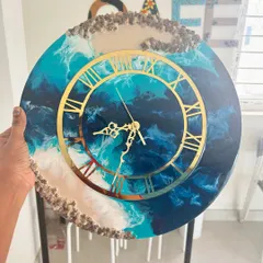 A close-up of a custom geode resin clock with stunning gold Roman numerals. The layers of blue and white resin mimic ocean waves, and the piece is finished with decorative stones.