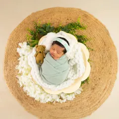 An overhead shot of a swaddled newborn in a flower-shaped basket, placed on a rustic jute mat with a tiny teddy bear prop.