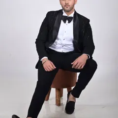 A model showcasing a classic black tuxedo with a modern twist. The jacket features subtle tonal embroidery on the shoulders and a satin shawl lapel, paired with a textured bow tie.