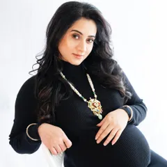 A modern and chic portrait of an expecting mother in a black turtleneck dress. She is seated and accessorized with a statement necklace, looking directly at the camera with confidence.