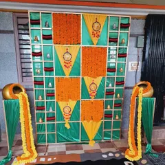 A traditional backdrop for a Bangle ceremony (Balesh Shastra). The design uses a grid pattern with panels of marigolds, fabric, and hanging bells, with decorative pots of overflowing flowers.