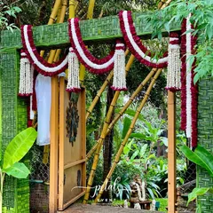 This traditional wedding entrance arch is wrapped in woven coconut leaves and decorated with garlands of maroon and white chrysanthemums, creating a rich, natural texture.