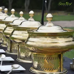 An outdoor buffet line with gleaming brass serving pots. I ensure my setups are not only functional but also aesthetically pleasing, complementing the decor of the event, whether it's indoors or outdoors.