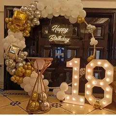 A glamorous 18th birthday entrance decor. This setup features a gold, silver, and white balloon arch over a doorway, a 'Happy Birthday' neon sign, and a large '18' marquee light.