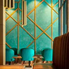A seating nook in the restaurant features plush teal velvet chairs set against a lime-washed wall with geometric brass inlay. The warm glow of the pendant lights creates an intimate and glamorous atmosphere.