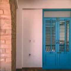 The vibrant blue of this louvered door creates a striking contrast against the earthy texture of the exposed CSEB wall.