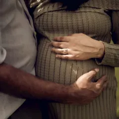 A warm, sunlit close-up of the couple's hands on the baby bump. The light and shadow create a beautiful texture, highlighting their gentle touch.
