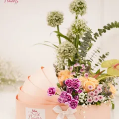 The 'Wild Grace' arrangement, designed in a unique cradle-shaped box. This piece features tall alliums, peach roses, purple carnations, and lush greenery for an elegant and unconventional look.