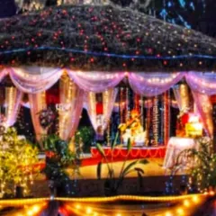 An outdoor Ganpati setup inside a gazebo structure. The hut-like roof and surrounding area are decorated with colorful string lights, creating a warm and inviting atmosphere for evening darshan.