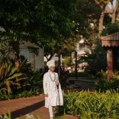 A full-length portrait of the groom in his elegant white and green sherwani, standing confidently in a lush garden setting.