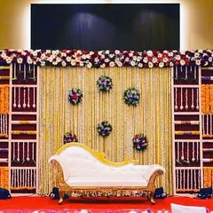 An elaborate stage decoration for a traditional ceremony. This setup uses a backdrop made of floral panels and shelving, with marigold garlands and a classic white chaise lounge for seating.