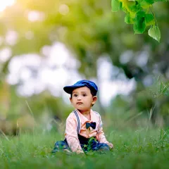 An outdoor portrait of a toddler boy dressed smartly with a cap and bow tie, sitting in the grass. The natural sunlight and green surroundings create a beautiful, soft-focus background.