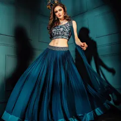 A full-length portrait of a model in a flowing teal lehenga. The studio lighting creates deep shadows that add dimension and mood, emphasizing the elegant silhouette of the outfit.
