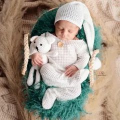 A sleeping newborn in a cozy, textured white romper and matching hat, holding a small knitted toy. This boho-inspired shot uses natural textures like pampas grass for a warm, earthy feel.