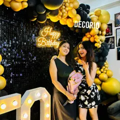 The birthday girl and her mother posing in front of a surprise party setup. The decor features a black sequin wall, a black and gold balloon arch, and a 'Happy Birthday' neon sign.
