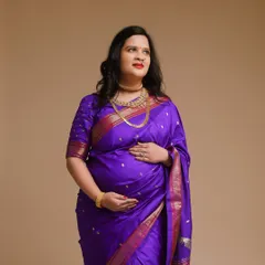 A classic studio portrait of a mom-to-be in a rich purple silk saree. The simple background and elegant pose highlight the beauty of the traditional attire.