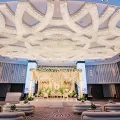 A wide view of a magnificent ballroom stage, designed with ivory and gold jharokhas and a ceiling draped in white floral garlands. This setup showcases a blend of royal Indian architecture and soft, romantic botanicals for a truly grand wedding.