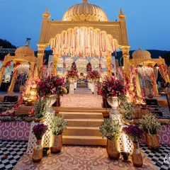 A beautiful twilight view of the main aisle leading to the mandap. The golden structures are illuminated, and the path is lined with lush florals and traditional vases, creating a majestic entrance.