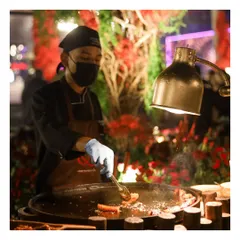 A chef at a live tawa station, illuminated by a heat lamp against a backdrop of vibrant red florals. This image captures the ambiance and professionalism of our outdoor catering setups, where every detail is considered.
