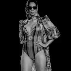 A powerful black and white shot of a model in a swimsuit and robe, styled with a headscarf and sunglasses. The dark background and strong pose create a look of timeless, cinematic glamour.