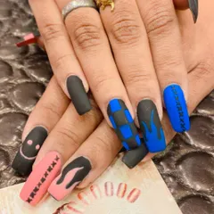 An edgy and eclectic set of long, matte nails. The design includes a mix of black, pink, and blue nails with hand-painted details like smiley faces, checkerboard patterns, and flame art.