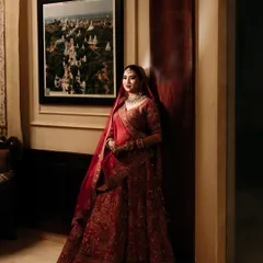 A classic bridal portrait showing the bride in her rich red lehenga. The indoor setting with warm lighting and a framed picture in the background adds a touch of sophistication.